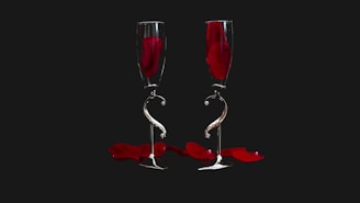 Two elegant champagne flutes are placed side by side against a dark background. Each glass contains a single red rose petal, and more petals are scattered below. The flutes have a unique twisting design on their stems, creating a sense of artistic sophistication.