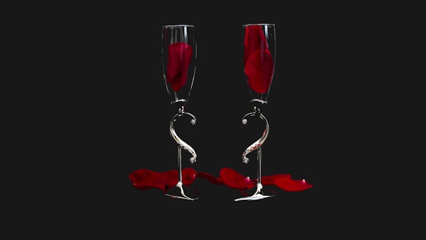 Two elegant champagne flutes are placed side by side against a dark background. Each glass contains a single red rose petal, and more petals are scattered below. The flutes have a unique twisting design on their stems, creating a sense of artistic sophistication.
