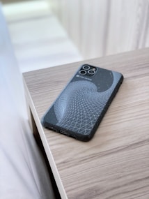 A smartphone with a unique geometric design on its case is placed on a light wooden surface. The case features a black background with a white swirling grid pattern, creating a modern and abstract look. The surface reflects a soft natural light, contributing to a clean and minimalist aesthetic.