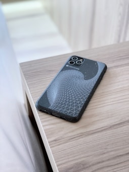 A smartphone with a unique geometric design on its case is placed on a light wooden surface. The case features a black background with a white swirling grid pattern, creating a modern and abstract look. The surface reflects a soft natural light, contributing to a clean and minimalist aesthetic.