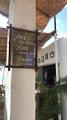 A rustic seaside cafe with a motivational sign that reads 'Live every moment, Love beyond words, Laugh everyday.' The setting includes whitewashed walls, a thatched roof, potted tropical plants, and the ocean visible in the background. There are framed pictures on the wall and various decorations contributing to a cozy and relaxed atmosphere.