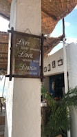 A rustic seaside cafe with a motivational sign that reads 'Live every moment, Love beyond words, Laugh everyday.' The setting includes whitewashed walls, a thatched roof, potted tropical plants, and the ocean visible in the background. There are framed pictures on the wall and various decorations contributing to a cozy and relaxed atmosphere.