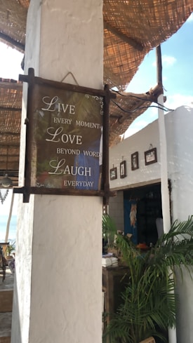 A rustic seaside cafe with a motivational sign that reads 'Live every moment, Love beyond words, Laugh everyday.' The setting includes whitewashed walls, a thatched roof, potted tropical plants, and the ocean visible in the background. There are framed pictures on the wall and various decorations contributing to a cozy and relaxed atmosphere.