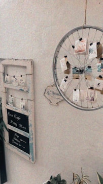 A rustic setup featuring a vintage-style decor element. On the left, there is a distressed wooden board with three chalkboard sections, labeled 'Eva Cafe', 'Happy Place', and 'Hope Believe Dream'. A bicycle wheel functions as a creative photo display, hanging on the wall with small pictures attached by clips. A small wooden sign with the word 'Beach' points towards the corner. Plants are partially visible at the bottom.