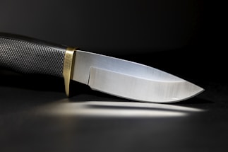 A sleek, sharp knife with a textured black handle and a shiny metallic blade. The knife has a brass bolster, and it is set against a dark background, casting a subtle reflection on the surface.