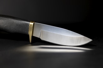 A sleek, sharp knife with a textured black handle and a shiny metallic blade. The knife has a brass bolster, and it is set against a dark background, casting a subtle reflection on the surface.