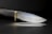 A sleek, sharp knife with a textured black handle and a shiny metallic blade. The knife has a brass bolster, and it is set against a dark background, casting a subtle reflection on the surface.