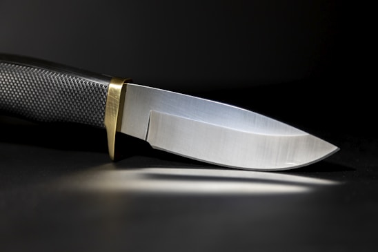 A sleek, sharp knife with a textured black handle and a shiny metallic blade. The knife has a brass bolster, and it is set against a dark background, casting a subtle reflection on the surface.