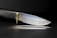 A sleek, sharp knife with a textured black handle and a shiny metallic blade. The knife has a brass bolster, and it is set against a dark background, casting a subtle reflection on the surface.