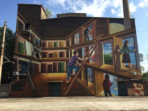 A large wall mural depicts a surreal scene with people interacting with books and climbing ladders. The painting uses a trompe-l'oeil effect, making the wall appear as shelves filled with oversized books, some of which are open like doors, revealing landscapes and figures stepping through them.