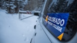 A vehicle is parked on a snowy path surrounded by trees. A sticker labeled 'Colorado Mountain College' is prominently displayed on the window of the car. In the background, a barrier blocks the path ahead.
