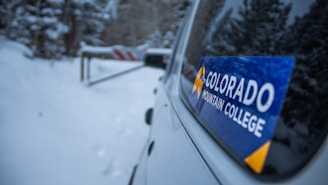 A vehicle is parked on a snowy path surrounded by trees. A sticker labeled 'Colorado Mountain College' is prominently displayed on the window of the car. In the background, a barrier blocks the path ahead.