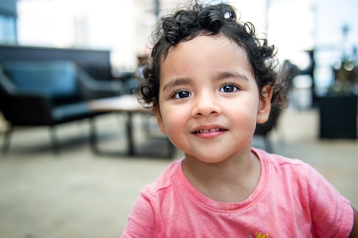 A young child with curly hair and a warm smile is wearing a pink shirt. The background appears to be a softly blurred indoor setting, possibly a living room or waiting area with dark furniture.