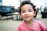 A young child with curly hair and a warm smile is wearing a pink shirt. The background appears to be a softly blurred indoor setting, possibly a living room or waiting area with dark furniture.