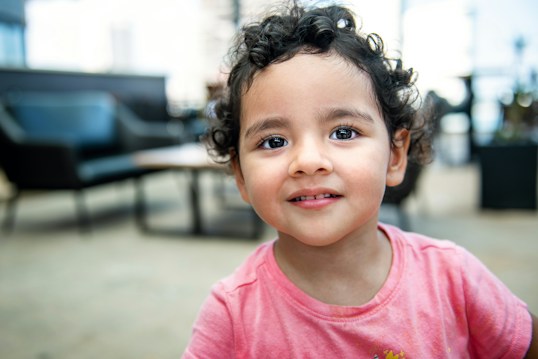 A young child with curly hair and a warm smile is wearing a pink shirt. The background appears to be a softly blurred indoor setting, possibly a living room or waiting area with dark furniture.