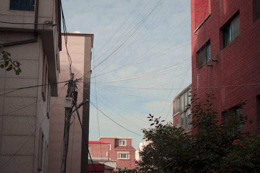 Urban alleyway between buildings with overhead wires converging at a utility pole. The buildings have a mix of red brick and beige facades. Some greenery is visible in the foreground, and the sky is partly cloudy.