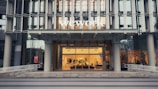 The entrance of a modern office building displays the large 'wework' logo above its door. The structure features a glass facade with a visible interior showing desks and office furniture. The exterior includes metal framework that adds to the contemporary aesthetic.