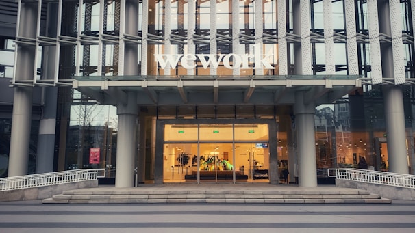 The entrance of a modern office building displays the large 'wework' logo above its door. The structure features a glass facade with a visible interior showing desks and office furniture. The exterior includes metal framework that adds to the contemporary aesthetic.