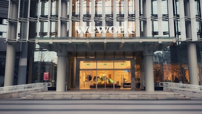 The entrance of a modern office building displays the large 'wework' logo above its door. The structure features a glass facade with a visible interior showing desks and office furniture. The exterior includes metal framework that adds to the contemporary aesthetic.