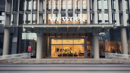 The entrance of a modern office building displays the large 'wework' logo above its door. The structure features a glass facade with a visible interior showing desks and office furniture. The exterior includes metal framework that adds to the contemporary aesthetic.