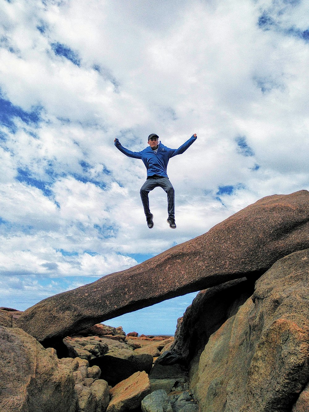 man in blue jacket jumping on brown rock formation during daytime