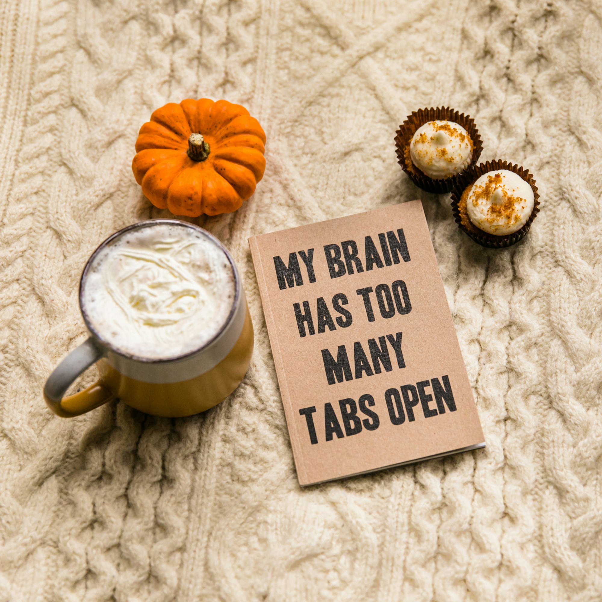 a sign saying "my brain has too many open tabs" is surrounded by a white ceramic mug with coffee, two cupcakes and a pumpkin