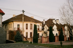 A suburban street scene featuring several houses with distinctive brown tiled roofs and cream-colored facades. The houses appear to have multiple stories with various architectural details such as balconies and chimneys. Tall, narrow evergreen trees are positioned along the property boundaries, and brick walls with black metal gates provide enclosure. Power lines cross overhead, and a partly cloudy sky casts soft, diffused lighting on the scene.