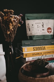 A decorative arrangement with dried flowers in a vase next to a stack of books. The books have colorful spines with titles related to recipes and vegetables. On top of the books is a bag labeled 'Father's,' featuring an illustration of a family cooking. The setting has a cozy and rustic feel.
