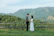 A happy couple standing in front of their new countryside lot with lush greenery around.