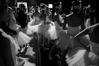Several ballerinas in tutus are gathered backstage, preparing for a performance. The dimly lit scene captures them from the side and behind, showing the delicate texture of their costumes and graceful postures as they chat and adjust their attire.