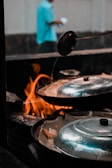 A cooking scene with a focus on pans covered with metal lids and a visible flame underneath. A person in a blurred background, wearing a blue shirt, is holding a white object.