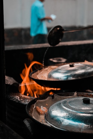 A cooking scene with a focus on pans covered with metal lids and a visible flame underneath. A person in a blurred background, wearing a blue shirt, is holding a white object.