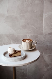 A round white table with a slice of cake topped with a scoop of vanilla ice cream on a plate, next to a cup of cappuccino with a heart-shaped latte art in a saucer. The background features a neutral tone marble wall.