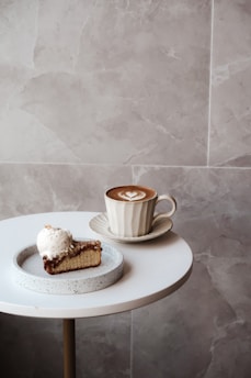 A round white table with a slice of cake topped with a scoop of vanilla ice cream on a plate, next to a cup of cappuccino with a heart-shaped latte art in a saucer. The background features a neutral tone marble wall.