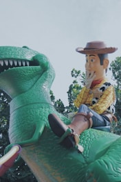 A large green dinosaur toy with a textured, scaly body is ridden by a cowboy toy wearing a brown hat, yellow checkered shirt, blue jeans, and brown boots. The cowboy displays a playful expression with one hand on his face. The scene is set against a backdrop of trees and a light blue sky.