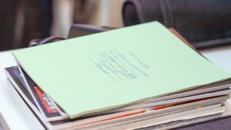 A stack of papers and notebooks is arranged on a table, with a green-covered book placed on top featuring handwritten text. In the background, a blurred black object is visible.