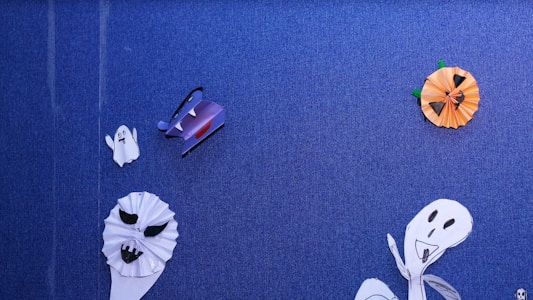 A blue textured background decorated with handmade Halloween-themed paper crafts. There are white paper ghosts with black eyes and mouths, a purple bat with wings, and an orange paper pumpkin with a jack-o'-lantern face. The crafts are simple and childlike, adding a playful and festive mood.