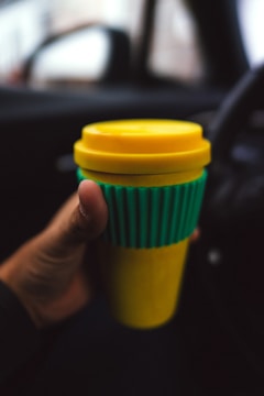 A hand holds a reusable coffee cup with a yellow lid and base, and a green ribbed midsection. The background is blurred, suggesting the setting is inside a car.