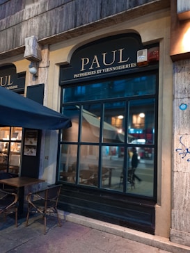 The storefront of a patisserie with a large window is visible. The shop has a dark exterior, with the name 'PAUL' and the words 'PATISSERIES ET VIENNOISERIES' written above the window. Outdoor seating includes a table and chairs under a navy blue awning. Inside the window, some light and faint reflections of the street can be seen.