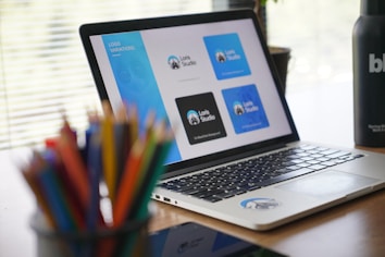 A laptop displaying various logo designs for 'Loris Studio' is placed on a wooden desk. The screen shows different blue and black logo variations. In the foreground, there is a blurry assortment of colored pencils. On the right side of the desk, a black bottle with white text is partially visible. Horizontal blinds allow natural light to softly illuminate the workspace.