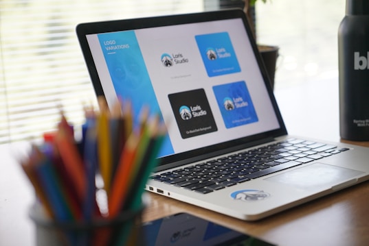 A laptop displaying various logo designs for 'Loris Studio' is placed on a wooden desk. The screen shows different blue and black logo variations. In the foreground, there is a blurry assortment of colored pencils. On the right side of the desk, a black bottle with white text is partially visible. Horizontal blinds allow natural light to softly illuminate the workspace.