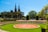 A sunny garden scene with well-manicured lawns and a circular decorative pattern in the center. The area is surrounded by neatly trimmed hedges, vibrant flower beds, and several tall street lamps. In the background, a historic building with pointed spires rises above the trees.