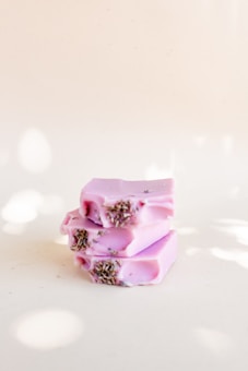 Three bars of lavender soap are stacked on each other. The soap is pastel purple in color, with dried lavender buds embedded on top. The background is a soft, light cream color.