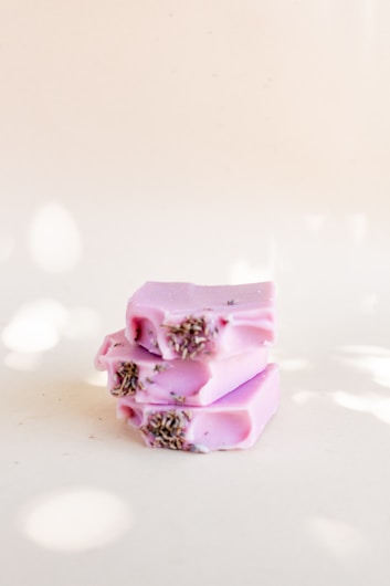 Three bars of lavender soap are stacked on each other. The soap is pastel purple in color, with dried lavender buds embedded on top. The background is a soft, light cream color.