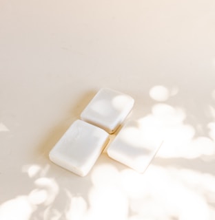 Three white soap bars are arranged on a light surface with soft shadows and light spots created by dappled sunlight.
