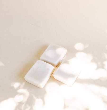 Three white soap bars are arranged on a light surface with soft shadows and light spots created by dappled sunlight.