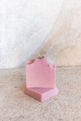 Two bars of pink soap, with one bar leaning against the other on a textured, light-colored surface. The soap has a smooth appearance with small specks, possibly of natural ingredients, and a rough edge on top.