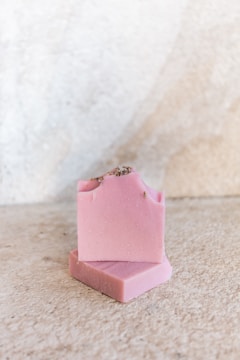 Two bars of pink soap, with one bar leaning against the other on a textured, light-colored surface. The soap has a smooth appearance with small specks, possibly of natural ingredients, and a rough edge on top.