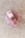A bar of pink soap is placed on a light textured surface. The soap has small dried herbs embedded on top, giving it a natural and artisanal appearance.