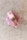 A bar of pink soap is placed on a light textured surface. The soap has small dried herbs embedded on top, giving it a natural and artisanal appearance.